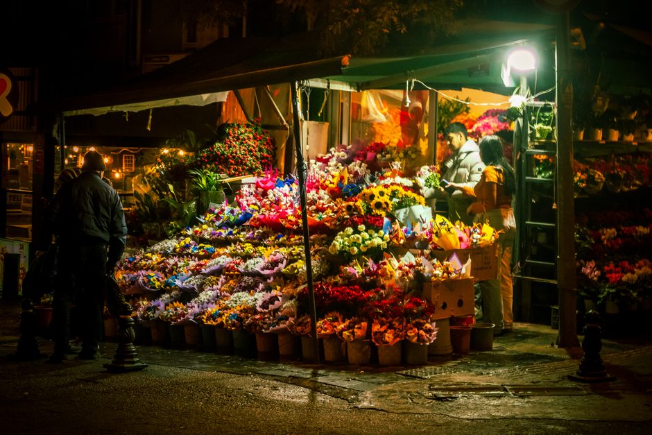 A vibrant flower stall at night illuminated by bright lighting, displaying an extensive selection of fresh, colorful flowers including sunflowers, roses, and daisies arranged in bouquets and bundles. 
