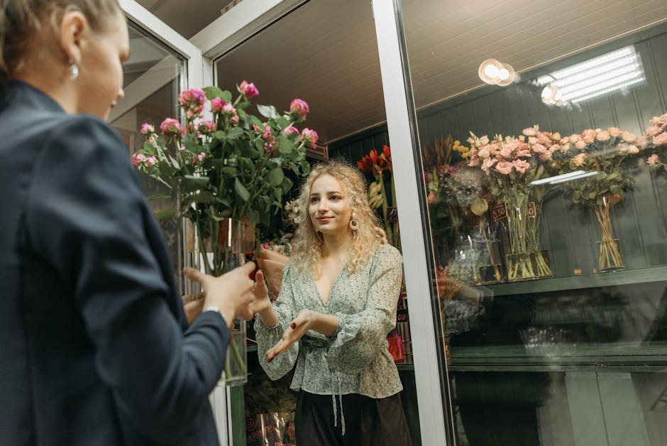 A female florist with curly blonde hair, dressed in a light patterned blouse, is engaged in a floral arrangement consultation at a flower shop. She is receiving a bouquets of pink roses with lush gree